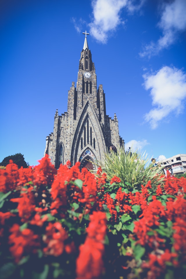 Picture of a Catholic church against a blue sky, surrounded by red flowers.